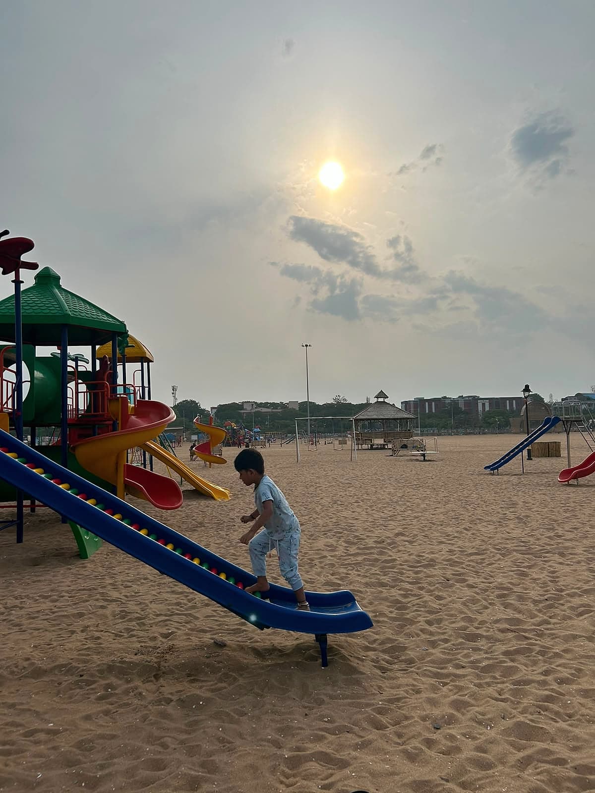 A small child climbing up a blue slide on a wide sandy playground, under a hazy sun.